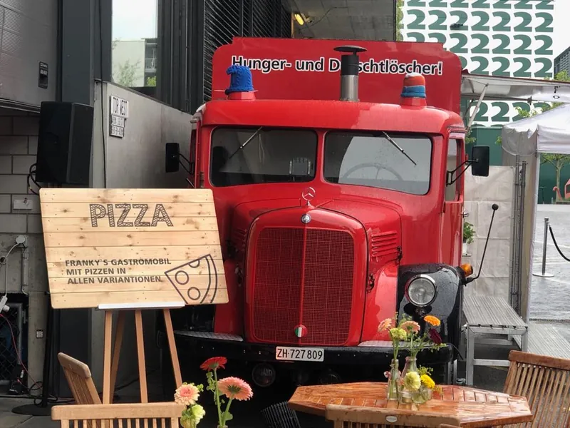 Das Feuerwehrauto von vorn, davor eine Holztafel mit der Aufschrift Pizza und Blumen auf den Tischen